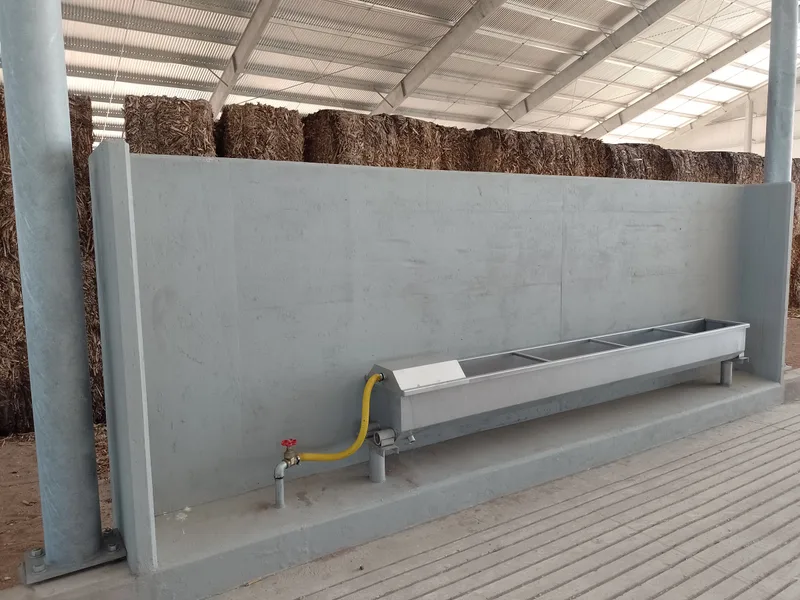 Tabique de hormigón armado con bebedero metálico y su bajada de agua, bajo la cubierta del corral y contra los fardos acopiados